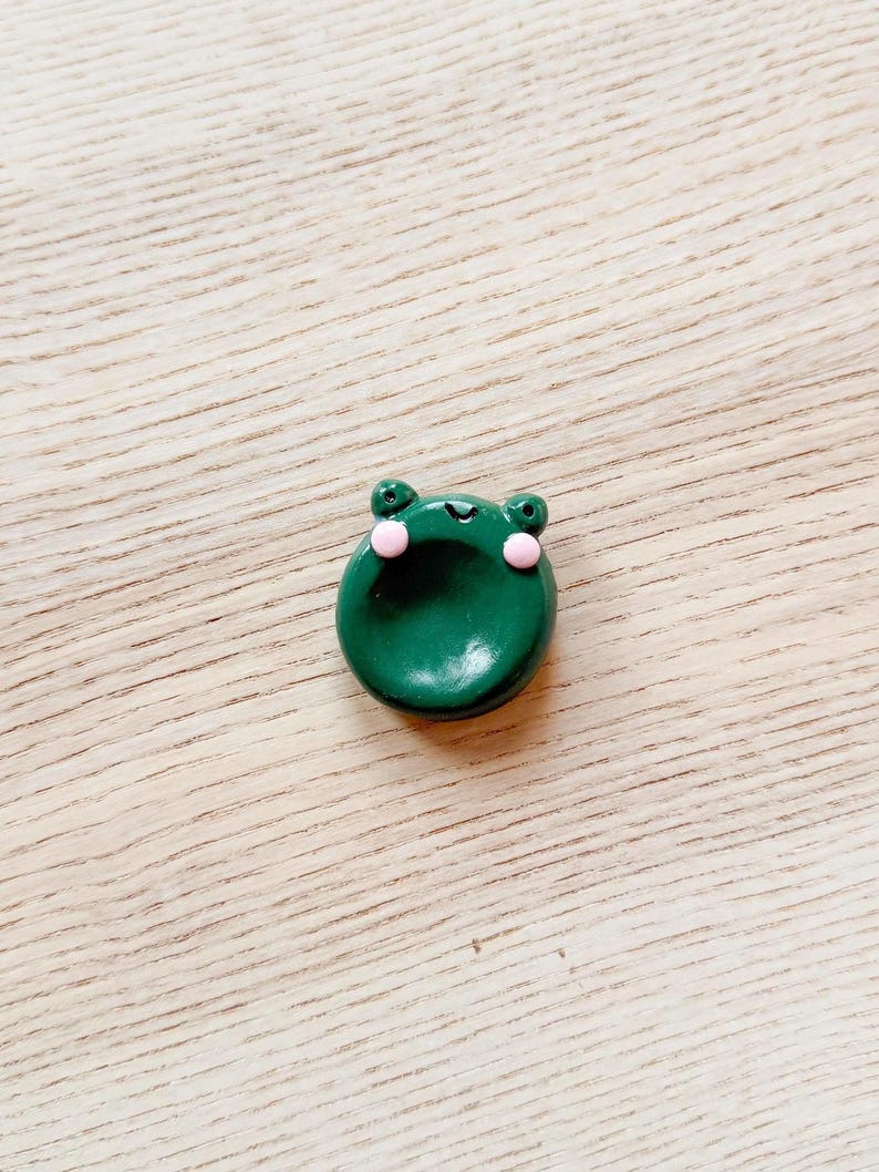 May include: A small, round, dark green frog-shaped object with pink cheeks and small, round eyes. The frog is sitting on a light brown wooden surface. The frog is a decorative item.