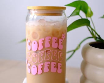 May include: A clear glass tumbler with a bamboo lid filled with iced coffee. The glass features the word COFFEE printed three times in a stacked, wavy pink font with a small pink flower icon next to the top word. The glass is filled with ice.