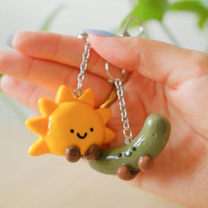 May include: A keychain featuring two small, glossy charms attached to silver-toned metal chains. One charm is a bright yellow sun with a smiling face and brown feet, and the other is a muted green pickle with a smiling face and brown feet.