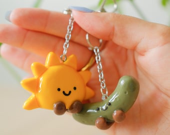 May include: A keychain featuring two small, glossy charms attached to silver-toned metal chains. One charm is a bright yellow sun with a smiling face and brown feet, and the other is a muted green pickle with a smiling face and brown feet.