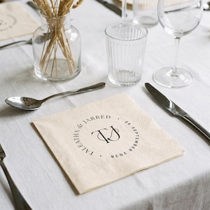 May include: A square, cream-colored paper napkin featuring a black circular monogram design. The text reads TALEATHA & JARRED and 26 SEPTEMBER 2026, surrounding a central TJ monogram. The napkin has a textured, embossed border pattern.