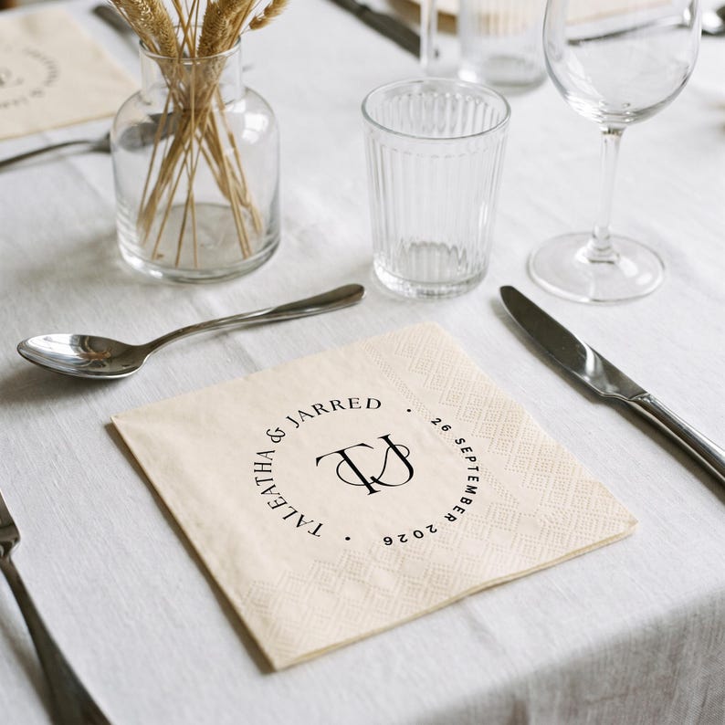 May include: A square, cream-colored paper napkin featuring a black circular monogram design. The text reads TALEATHA & JARRED and 26 SEPTEMBER 2026, surrounding a central TJ monogram. The napkin has a textured, embossed border pattern.