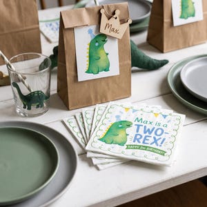 May include: A brown paper gift bag with a wooden crown tag labeled Max, featuring a green dinosaur illustration. Beside it, a clear glass with a dinosaur print and a stack of white napkins printed with a green dinosaur and the text Max is a TWO-REX!