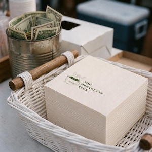 May include: A stack of square, light-colored paper napkins featuring a green graphic of a coffee cup and the text THE BREAKFAST CLUB, placed inside a white woven basket with a wooden handle, next to a metal container filled with US dollar bills.