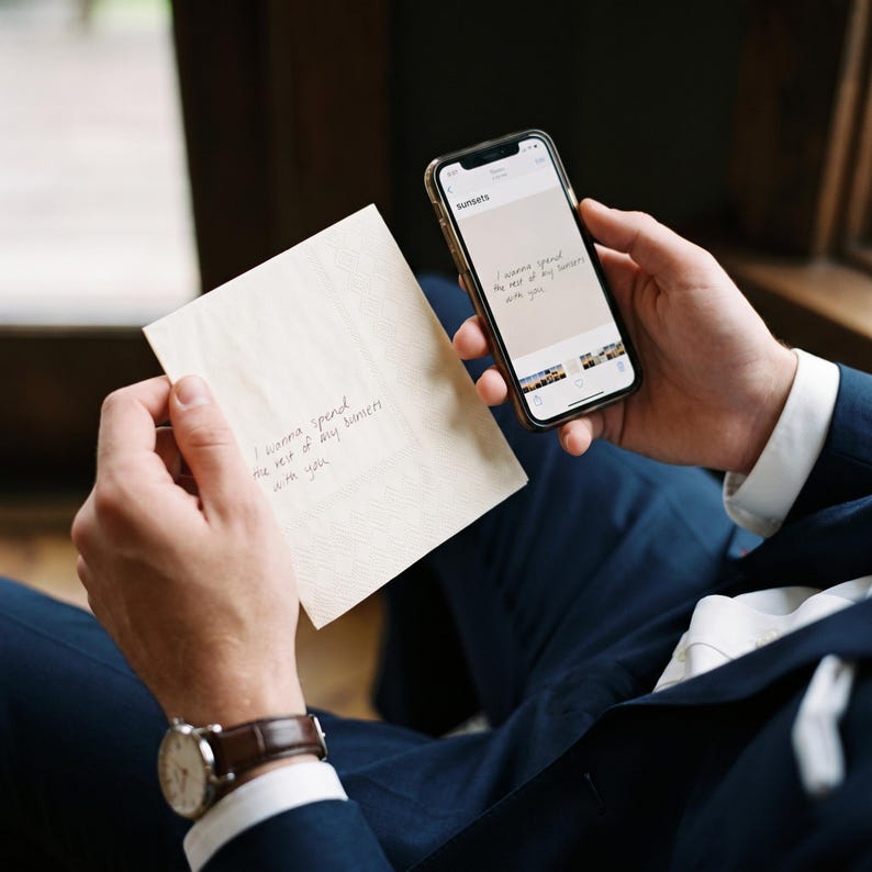 May include: A cream-colored square card with a textured border and the handwritten text "I wanna spend the rest of my sunsets with you" held alongside a smartphone displaying the same text on its screen, both held by a person in a navy suit.