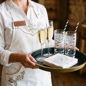 May include: A round black serving tray with a gold rim holding two tall champagne flutes filled with sparkling liquid, two highball glasses with ice and black-and-white striped straws, and a stack of white napkins printed with Cowgirl Cocktail Club.