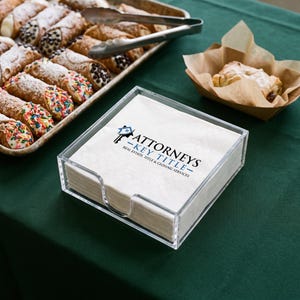 May include: A stack of white paper napkins held in a clear acrylic square holder. The top napkin features a printed logo with a house icon, a key graphic, and the text Attorneys Key Title Real Estate, Title & Closing Services in black and blue.