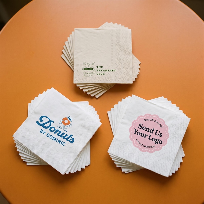 May include: Three stacks of square white paper napkins. The top napkin of the first stack features a green coffee cup graphic and the text THE BREAKFAST CLUB. The second stack shows a blue donut character and the text DONUTS BY DOMINIC. The third stack displays a pink scalloped circle with the text SEND US YOUR LOGO.