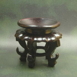 May include: A dark brown wooden plant stand with a round top and intricate carvings. The stand has three legs with curved designs.