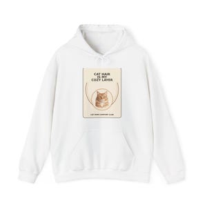 May include: A white hooded sweatshirt featuring a rectangular graphic on the chest. The graphic contains a beige background, a circular frame, an illustration of an orange tabby cat, and the text CAT HAIR IS MY COZY LAYER and CAT MOM COMFORT CLUB.