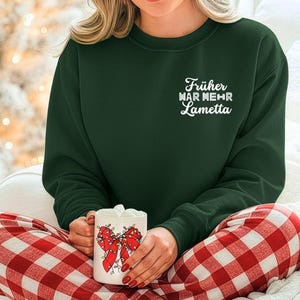 Könnte beinhalten: Ein dunkelgrünes Sweatshirt mit dem weißen Schriftzug "Früher war mehr Lametta". Die Person trägt eine rot-weiß karierte Pyjamahose und hält einen weißen Becher mit einem roten Schleifendesign.