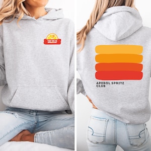 Pode incluir: Um moletom cinza claro com bolso frontal e capuz. A frente apresenta um pequeno gráfico de uma fatia de limão e o texto "THEY SEE ME APEROLIN'". A parte de trás exibe listras horizontais laranja e vermelha com o texto "APEROL SPRITZ CLUB".