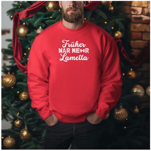 Könnte beinhalten: Ein rotes Sweatshirt mit dem weißen Text "Früher war mehr Lametta". Das Sweatshirt ist einfarbig und hat einen Rundhalsausschnitt. Das Bild zeigt eine Person, die das Sweatshirt vor einem Weihnachtsbaum trägt.