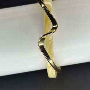 May include: A gold-toned bangle bracelet with a wavy design. The bracelet is made of a white material with a marbled pattern.