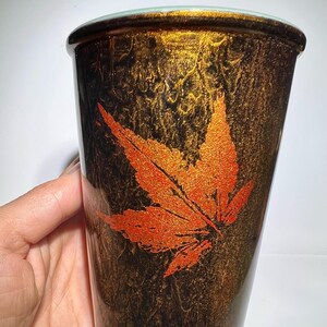 May include: A tall, cylindrical tumbler with a textured, dark brown and gold finish. An orange maple leaf design is prominently displayed on the side. The rim is a light green color.