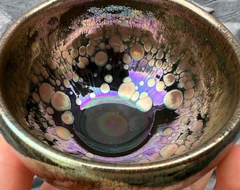 Könnte beinhalten: Eine kleine, dunkelbraune Teetasse aus Keramik mit einer strukturierten, irisierenden Glasur. Die Glasur hat ein Muster aus kleinen, runden Blasen.