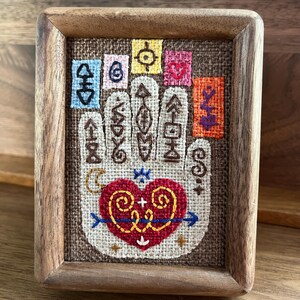 May include: A framed embroidery depicting a hand with a heart and symbols in a range of colours, including red, yellow, blue, and green. The hand is surrounded by small squares with different symbols and colours.
