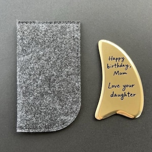 May include: A gold-colored gua sha tool with the inscription "Happy birthday, Mom, Love your daughter" next to a gray felt pouch. The tool has a smooth, curved shape. The pouch is rectangular with rounded corners.
