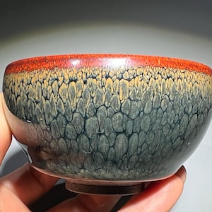 Può includere: Una piccola ciotola in ceramica verde scuro con una glassa testurizzata e macchiata. La ciotola ha un bordo rosso e un fondo leggermente arrotondato.