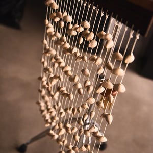 Puede incluir: Un instrumento musical hecho de un marco de madera con múltiples cuerdas que sostienen pequeños objetos de color marrón claro con forma de concha. El instrumento está montado en un soporte de metal.