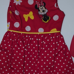 Puede incluir: Vestido rojo sin mangas con lunares blancos y ribete amarillo. El corpi&ntilde;o presenta un gr&aacute;fico de Minnie Mouse, margaritas blancas y lazos amarillos. La falda est&aacute; fruncida y tiene un estampado de lunares blancos.