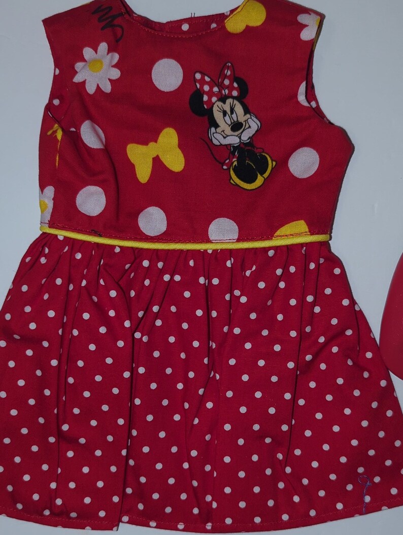 Puede incluir: Vestido rojo sin mangas con lunares blancos y ribete amarillo. El corpi&ntilde;o presenta un gr&aacute;fico de Minnie Mouse, margaritas blancas y lazos amarillos. La falda est&aacute; fruncida y tiene un estampado de lunares blancos.