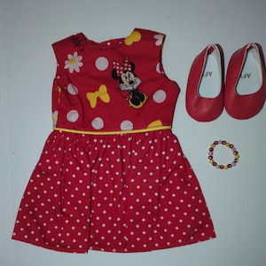 Puede incluir: Un vestido de mu&ntilde;eca rojo con lunares blancos, un lazo amarillo y un gr&aacute;fico de Minnie Mouse. El conjunto incluye zapatos de mu&ntilde;eca rojos y una pulsera de cuentas. El vestido tiene un ribete amarillo.