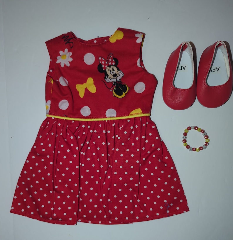 Puede incluir: Un vestido de mu&ntilde;eca rojo con lunares blancos, un lazo amarillo y un gr&aacute;fico de Minnie Mouse. El conjunto incluye zapatos de mu&ntilde;eca rojos y una pulsera de cuentas. El vestido tiene un ribete amarillo.