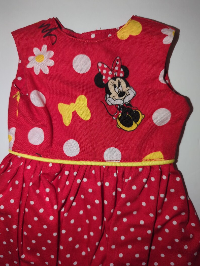 Puede incluir: Vestido rojo sin mangas con lunares blancos y ribete amarillo. El vestido presenta una imagen de Minnie Mouse, margaritas blancas y lazos amarillos. La falda est&aacute; fruncida y el vestido es de tela ligera.
