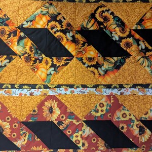 May include: A quilted textile featuring a repeating pattern of sunflowers and pumpkins in shades of yellow, orange, and black. The design incorporates geometric shapes and a border with autumn-themed motifs, creating a seasonal aesthetic.