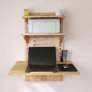 May include: A wall-mounted wooden desk with two shelves. The top shelf holds a printer and a mug. The middle shelf is open. The bottom shelf is a desk with a laptop, phone, mouse, and mousepad. The desk is made of light-colored wood.