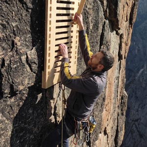 May include: A wooden climbing training board with horizontal slots is mounted on a rock face. A person is using the board, holding a wooden grip. The board is attached to the rock with a metal bracket. The person is wearing climbing gear.