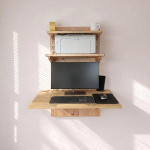 May include: A light-colored wooden wall-mounted desk with a monitor, keyboard, mouse, and printer. The desk has a small drawer and a shelf above it with a mug and tape roll. The desk is mounted on a light pink wall.