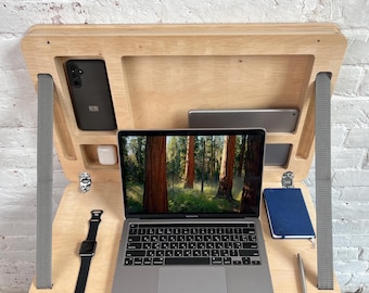 May include: A light-colored wooden laptop desk with built-in compartments for a phone, tablet, and accessories. A laptop, watch, notebook, and pen are on the desk. The laptop screen displays a forest scene. Gray straps support the desk.