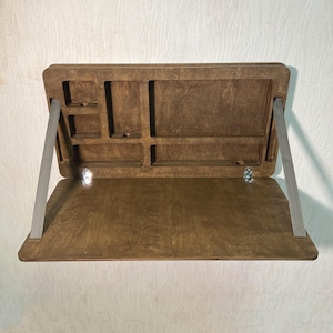 May include: A wooden, foldable laptop desk with a natural wood finish. The desk has a compartmented top section for storage and is supported by grey fabric straps. The desk is designed to be mounted on a wall.