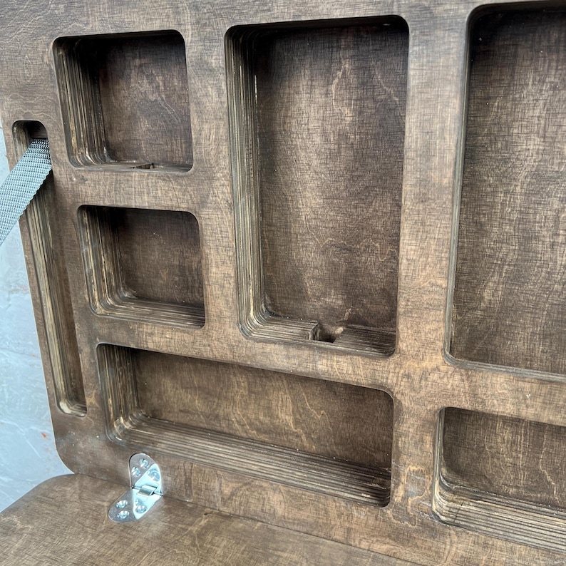 May include: A dark stained wooden organiser with a hinged lid and multiple compartments. The interior features various rectangular and square cutouts. A grey strap is attached to the side. A metal hinge is visible. The wood has a dark brown stain.