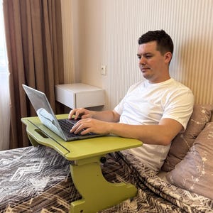 May include: A light green wooden laptop desk with adjustable height settings. The desk is placed on a bed with a patterned blanket. A person is using a laptop on the desk.