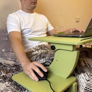 May include: A light green adjustable laptop desk with a black computer mouse. The desk has a top surface for a laptop and a lower surface for the mouse. The desk is supported by a curved, vertical support.