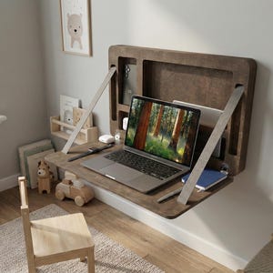May include: A dark wood, wall-mounted desk with a laptop showing a forest scene. The desk has a built-in storage compartment and is supported by two angled metal supports. A small wooden chair and toy car are in front of the desk.
