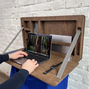 Puede incluir: Un escritorio de madera montado en la pared con un portátil, una tableta y un smartphone. El escritorio es marrón con compartimentos de almacenamiento y correas grises. Una persona está usando el portátil.