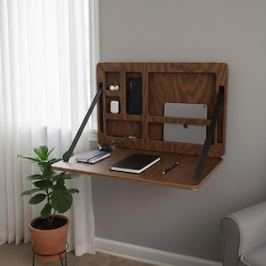 Può includere: Una scrivania in legno da parete con un design pieghevole. La scrivania è di colore marrone caldo e presenta scomparti per riporre un tablet, un telefono e altri piccoli oggetti. Sono visibili cinghie di supporto nere. Una pianta in vaso di terracotta è sotto.
