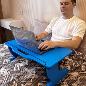 May include: A bright blue wooden laptop desk with angled legs. The desk is designed to sit on a bed or other surface, providing a stable workspace. The laptop is open and in use. The desk is a vibrant accent piece.