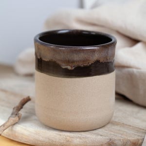 Può includere: Una tazza in ceramica fatta a mano con un design bicolore. La parte superiore è marrone scuro, che sfuma in una fascia marrone più chiara e poi in una base beige. La tazza è cilindrica e sembra fatta a mano.