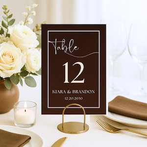May include: A brown table number card with the word "Table" in cursive script and the number "12" in a bold font. The card also includes the names "Kiara & Brandon" and the date "12/20/2030". The card is displayed in a gold metal holder.