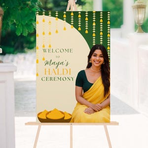 May include: A welcome sign for Maya's Haldi Ceremony. The sign features a woman in a yellow sari, with text in green and gold. The sign is on an easel, with a green and gold background.