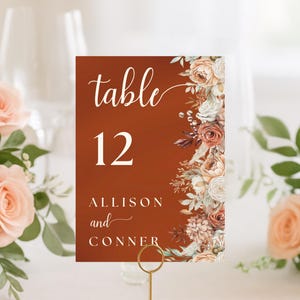 May include: A rectangular table number card with a burnt orange background, featuring the text table 12, ALLISON and CONNER, and a vertical floral arrangement of roses and foliage in shades of cream, peach, and rust along the right edge.