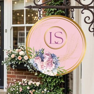 Puede incluir: Un letrero de tienda redondo rosa con borde dorado y las iniciales "LS" en un círculo central. El letrero presenta un arreglo floral de flores rosas y azules y está colgado de un soporte metálico decorativo.