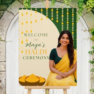 May include: A welcome sign for Maya's Haldi Ceremony. The sign features a woman in a yellow sari, with text that reads "Welcome to Maya's Haldi Ceremony." The sign has a cream background with yellow and green accents.