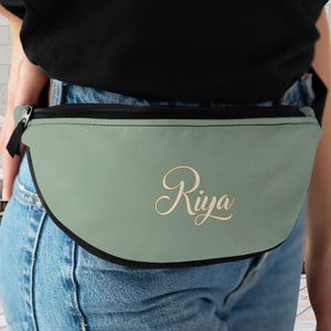 Könnte beinhalten: Eine salbeigrüne Bauchtasche mit schwarzem Reißverschluss und Besatz. Das Wort "Riya" ist in einer Schreibschrift auf der Vorderseite der Tasche geschrieben. Die Bauchtasche wird um die Taille getragen.