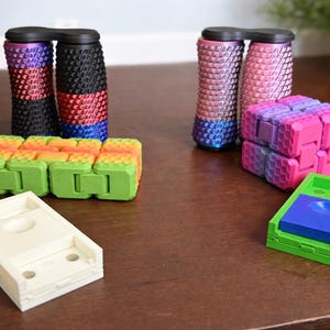 May include: An assortment of colorful fidget toys, including two sets of connected cylinders with textured surfaces, a rainbow-colored articulated block, a pink and purple cube, and a green and blue tray. The toys are arranged on a brown surface.
